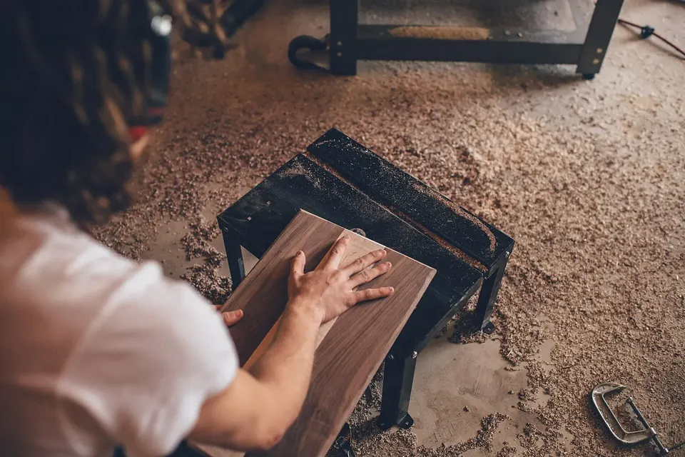 Artisan ponçant à la main une planche de bois sur un établi, dans un atelier au sol couvert de sciure