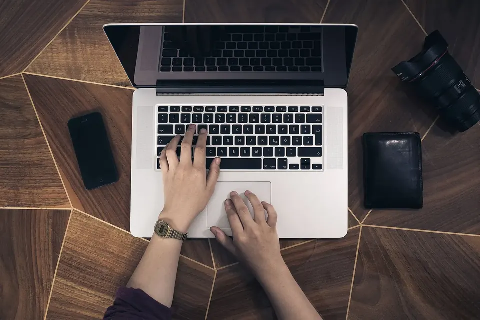 Deux mains tapant sur le clavier d'un ordinateur portable posé sur une table en bois, à côté d'un téléphone et d'un portefeuille