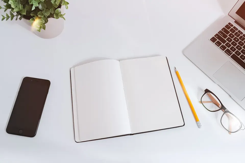 Table blanche avec un carnet ouvert, un crayon, des lunettes, un téléphone et un ordinateur portable